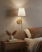 Gold wall sconce with a ribbed white fabric shade, lit in a warm bedroom setting - Silver