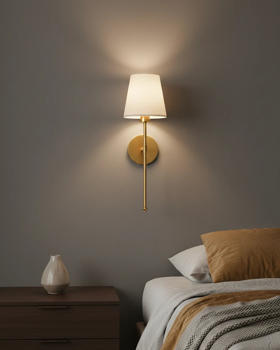 Gold wall sconce with a ribbed white fabric shade, lit in a warm bedroom setting