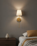 Gold wall sconce with a ribbed white fabric shade, lit in a warm bedroom setting