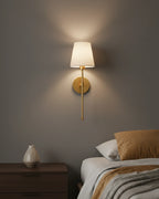 Gold wall sconce with a ribbed white fabric shade, lit in a warm bedroom setting
