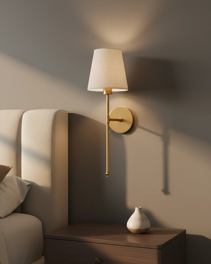 Gold wall sconce with a ribbed white fabric shade, lit in a warm bedroom setting