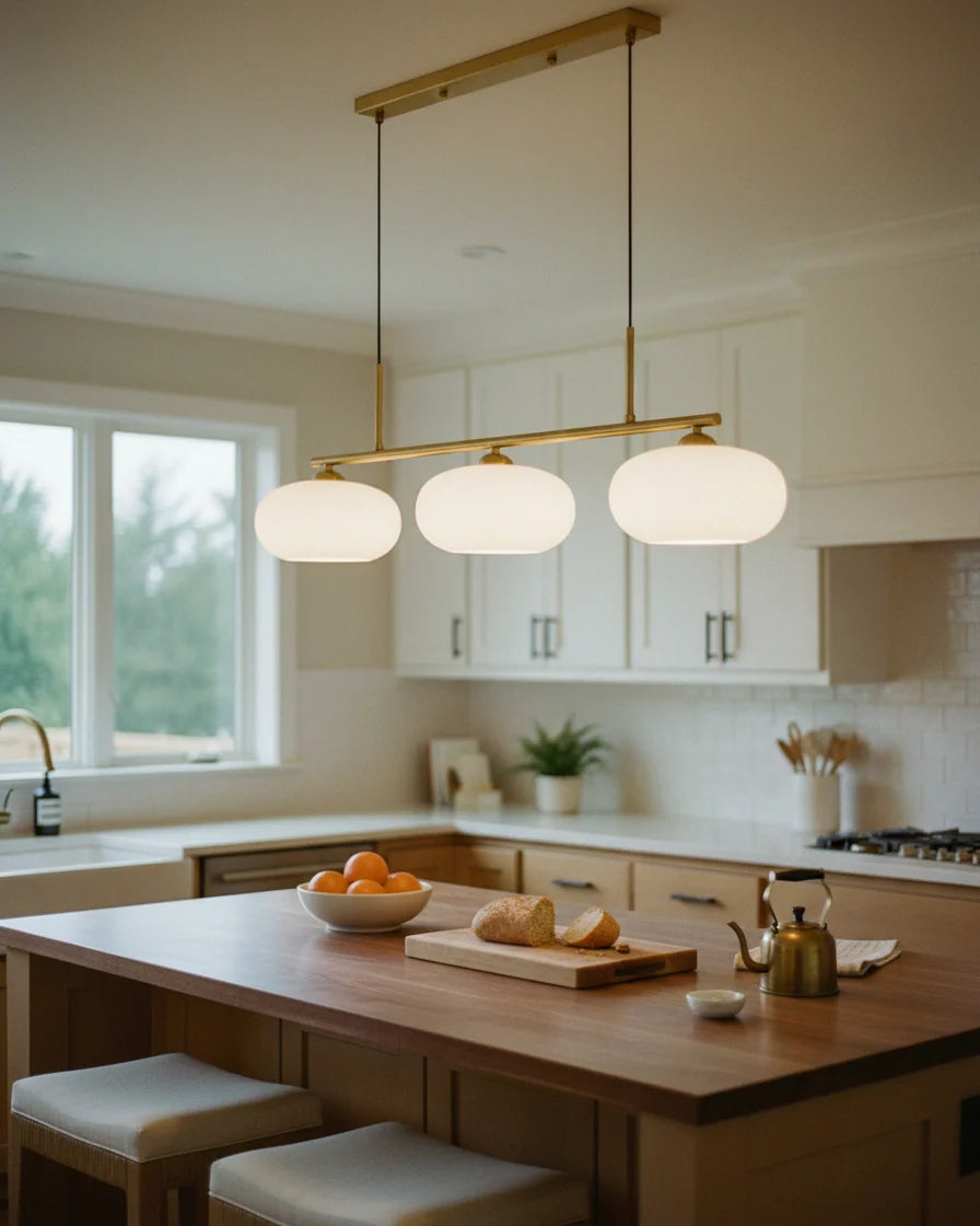 Linear 3-light pendant with gold finish and three spherical white glass shades, lit, in a kitchen setting - hero 3